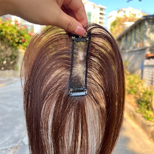 May include: A brown and blonde hairpiece with a black clip. The hairpiece is designed to add volume or length to existing hair. The clip is made of metal and fabric, and is attached to the hairpiece.