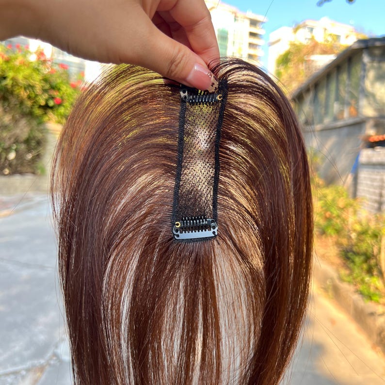 May include: A brown and blonde hairpiece with a black clip. The hairpiece is designed to add volume or length to existing hair. The clip is made of metal and fabric, and is attached to the hairpiece.