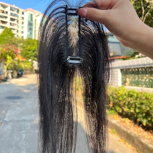 May include: A black hair topper with two metal clips. The hairpiece is held up against a background of green foliage and a building.