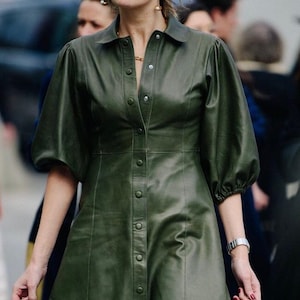Puede incluir: Un vestido de cuero verde oliva con botones en la parte delantera y mangas cortas abullonadas. El vestido tiene cuello camisero y cintura entallada. La persona lleva gafas de sol y un reloj. El vestido llega a la rodilla.