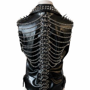 Op de afbeelding: Een zwart mouwloos vest met een kraag versierd met zilverkleurige metalen spikes. Het achterpand is gedecoreerd met een verticale rij zilveren veiligheidsspelden die verbonden zijn door meerdere gedrapeerde zilveren kettingen.