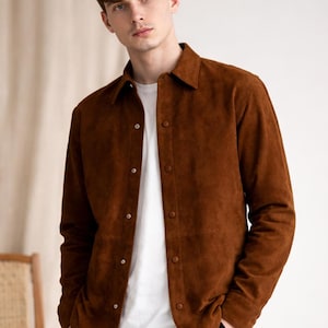 May include: A brown suede jacket with a collared neckline and button-up closure. The jacket is worn over a white t-shirt and paired with black pants. The model is standing in front of a neutral background.