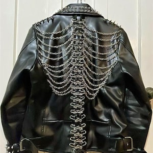 May include: A black faux leather jacket featuring silver-toned metal spikes along the collar and shoulders. The back is decorated with multiple draped silver chains and a vertical row of silver safety pins, with adjustable buckled straps at the waist.