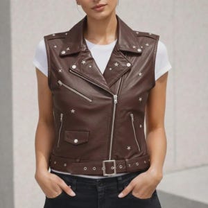 Op de afbeelding: Een bruin leren vest met zilveren sterren, een reverskraag en een diagonale ritssluiting. Het vest heeft een riem met een zilveren gesp en een klein zakje. Er wordt een wit t-shirt onder gedragen.