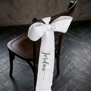 May include: A dark wooden chair with a white fabric bow tied to the back. The fabric has the name "Jordan" printed in a cursive font. The chair is set against a dark background.