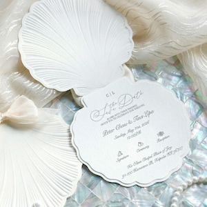 May include: A white, shell-shaped card with embossed ridges and scalloped edges. The text reads: C|L, Save the Date, Peter Chaw & Tina Lipa, Sunday, May 31st, 2026, 12:00PM, along with icons for Signature, Ceremony, and Reception at Ko Olina Chapel Place of Joy.