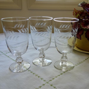 May include: Three clear stemmed wine glasses with etched wheat stalk designs. The glasses are arranged on a white tablecloth with a floral pattern. A potted plant with red and green leaves is in the background.