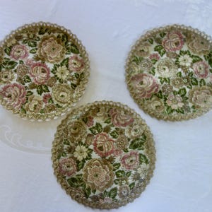 May include: Three circular decorative plates featuring a detailed floral tapestry pattern with pink, white, and green flowers on a cream background, finished with a scalloped, metallic gold-toned lace-like border around the edges.