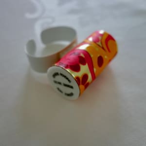 Puede incluir: Un recipiente cilíndrico con un colorido estampado floral en tonos rojos, naranjas y amarillos, junto a un anillo blanco y curvado. El recipiente tiene ribete dorado y el texto "CELINE PARIS".