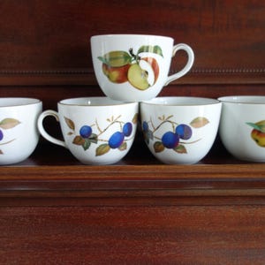 Puede incluir: Un conjunto de tazas y cuencos de cerámica blanca con bordes dorados. Las tazas y los cuencos están decorados con ilustraciones de frutas coloridas, como manzanas y ciruelas. Las tazas tienen asas, mientras que los cuencos no.