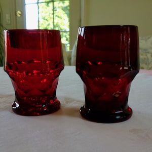 May include: Two ruby red drinking glasses with a faceted design. The glasses have a short, rounded base and a slightly flared top. They are sitting on a white tablecloth.
