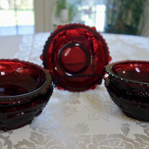 Puede incluir: Cuencos de vidrio rojo rubí y un centro de mesa decorativo. Los cuencos tienen un patrón texturizado y bordes festoneados. El centro de mesa tiene un diseño circular con un patrón texturizado. Los artículos están colocados sobre un mantel blanco.