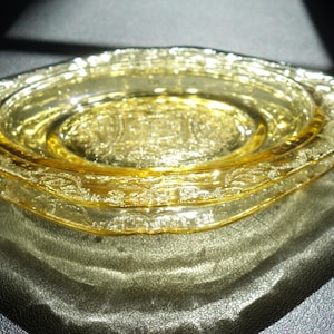 May include: A stack of three vintage, yellow-tinted glass dishes. The dishes have a decorative, embossed floral pattern around the edges. The light catches the glass, creating highlights and shadows.