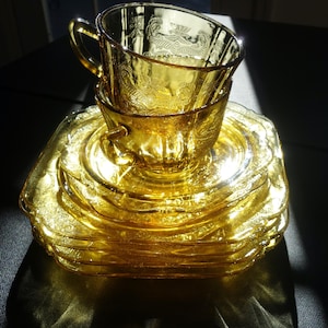 May include: A stack of vintage amber-colored glass teacups and saucers. The cups and saucers have a detailed embossed floral pattern. The set is illuminated by sunlight, creating a warm, inviting glow. The teacups are stacked on top of the saucers.