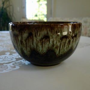 May include: A ceramic bowl with a glossy finish. The bowl has a dark brown exterior that fades into a green and brown pattern. The bowl is sitting on a white tablecloth.