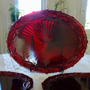 May include: A set of three ruby red glass plates. The large plate is round with a scalloped edge and a sunburst design in the center. The plates are displayed on a white tablecloth.