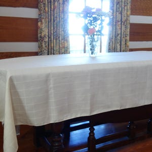 May include: A cream-colored rectangular tablecloth with a subtle grid pattern covers a dark wooden table. The table is set in front of a window with floral curtains and a vase of flowers. The room has wooden walls.