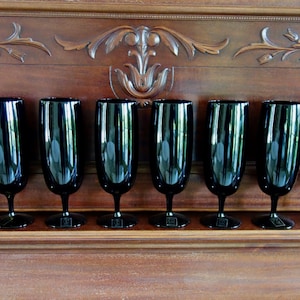 May include: A set of eight black champagne flutes with a sleek, modern design. The glasses are made of a dark, reflective material and stand on a wooden surface. The background features ornate wooden carvings.