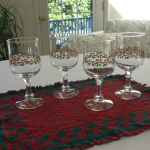 May include: Four clear wine glasses with gold rims, decorated with a festive red and green holly pattern. They are arranged on a red and green patterned placemat, creating a holiday theme.