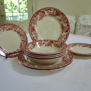 Op de afbeelding: Een vintage servies met een crèmekleurige basis en een rood bloemmotief. De set bevat borden, kommen en schotels, gerangschikt op een wit tafelkleed. De borden hebben een diameter van ongeveer 25 cm.