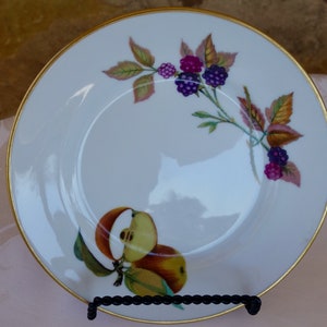 Puede incluir: Plato de porcelana blanca con borde dorado, con un diseño pintado de moras y manzanas. Las frutas y hojas son en tonos rojos, morados y verdes. El plato se exhibe sobre un soporte de metal negro.