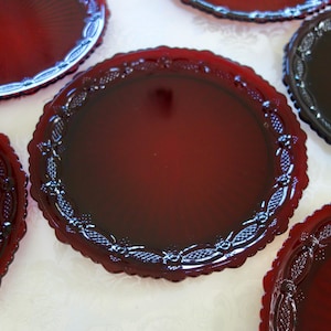 May include: A collection of ruby red glass plates with ornate, scalloped edges. The plates have a glossy finish and a detailed floral and leaf pattern around the rim. The plates are arranged on a light-colored surface.