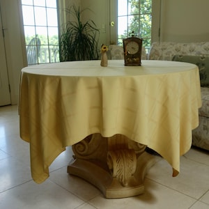 May include: A round, pale yellow tablecloth with a subtle, tonal checkered pattern draped over a circular table. The table features a decorative, carved pedestal base. A small clock and a tiny vase are placed on the center of the tabletop.