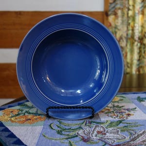 Puede incluir: Un cuenco de cerámica azul cobalto con anillos concéntricos blancos, exhibido sobre un soporte decorativo. El cuenco está sobre un mantel con estampado floral. El fondo incluye una pared de madera y una cortina floral.
