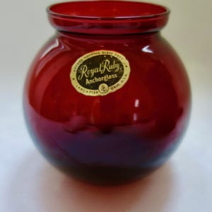 May include: A red glass vase with a round body and a wide mouth. The vase has a gold label that reads "Royal Ruby Anchorhoglass Lancaster Ohio U.S.A."