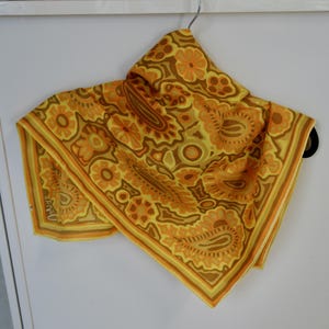 May include: A vintage scarf with a floral paisley pattern in shades of yellow, orange, and brown. The scarf is square-shaped and appears to be made of a lightweight fabric, hanging from a hanger.