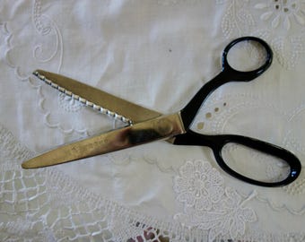 Könnte beinhalten: Vintage Schere mit schwarzen Griffen und silbernen Klingen, eine davon gezahnt. Die Schere liegt geöffnet auf weißem, besticktem Stoff. Auf den Klingen ist das Wort "ROBO" eingraviert. Der Stoff hat ein zartes Spitzenmuster.