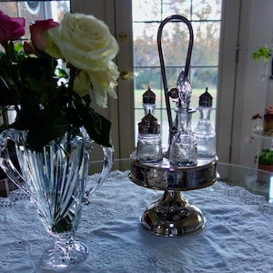 Puede incluir: Un jarrón de cristal contiene rosas en tonos rosa y blanco. Un juego de condimentos plateado con botellas y saleros de vidrio se encuentra sobre una base redonda. El juego tiene un asa y está sobre un mantel de encaje blanco.