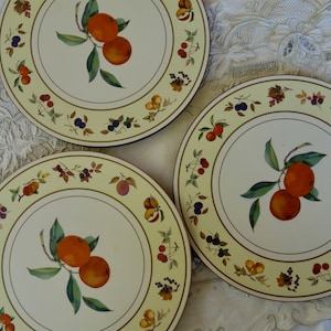 Puede incluir: Tres manteles individuales redondos, amarillos y blancos con un borde dorado. Cada mantel individual presenta una ilustración estilizada de una rama con fruta naranja y hojas verdes. Los manteles individuales están rodeados por un borde de pequeñas frutas y bayas de colores.