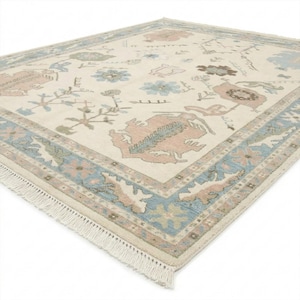 May include: Cream-colored area rug with a detailed floral pattern in shades of pink, blue, and green. The rug features a border with a repeating geometric design and fringe along the edge. The rug is rectangular.