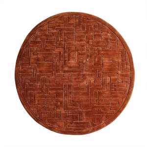 May include: A round, rust-colored rug with a geometric pattern. The rug features a series of interconnected rectangular shapes, creating a maze-like design. The rug is made of a soft material and has a textured surface.