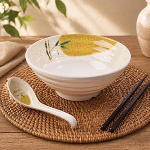 Peut inclure: Un bol en céramique blanc avec un motif de bambou jaune et vert, une cuillère assortie et des baguettes noires reposent sur un dessous de plat tissé. Le bol a un large bord et un extérieur texturé. La cuillère et le bol ont un motif de bambou.