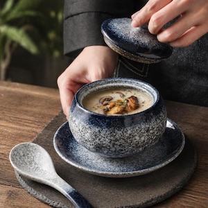 Peut inclure: Un bol à soupe en céramique avec un couvercle, rempli de soupe et garni de champignons. Le bol, la soucoupe et la cuillère présentent un motif moucheté bleu et blanc. Le couvercle est soulevé, révélant la soupe à l'intérieur.