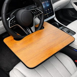 May include: A wooden car steering wheel desk with a black border is attached to a black steering wheel. The desk is light brown with a wood grain pattern. The car interior is white and black.