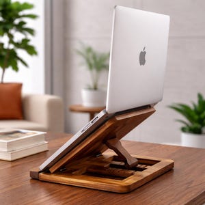 Könnte beinhalten: Ein Laptop-Ständer aus Holz mit einem silbernen Laptop. Der Ständer besteht aus hellbraunem Holz und hat eine rechteckige Basis mit einer erhöhten Plattform zur Aufnahme des Laptops. Der Laptop ist nach oben abgewinkelt.