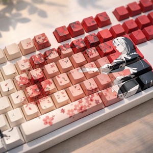 May include: A mechanical keyboard with custom keycaps. The keycaps feature a gradient of colors from white to red, with floral and anime-style artwork. The keyboard has a white frame and is set on a light-colored surface.