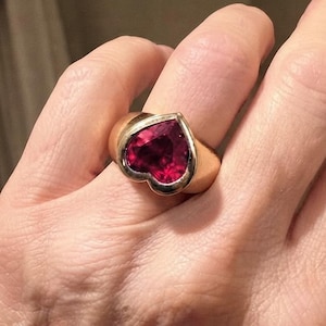 Pode incluir: Um anel de ouro com uma grande pedra preciosa em forma de coração de cor rosa intenso, engastada numa banda de ouro lisa e polida. O engaste segue a forma de coração da pedra, criando um ponto focal arrojado e proeminente no dedo.