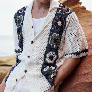 Può includere: Camicia all'uncinetto color crema con un motivo floreale nei toni del verde, blu navy e bordeaux. La camicia ha maniche corte, colletto e chiusura con bottoni. Il motivo all'uncinetto è aperto, creando un aspetto strutturato.