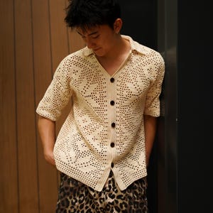 Op de afbeelding: Crèmekleurig, open geweven gehaakt overhemd met korte mouwen en een kraag. Het overhemd heeft donkerbruine knopen en wordt gecombineerd met een short met luipaardprint. De outfit is casual en stijlvol.