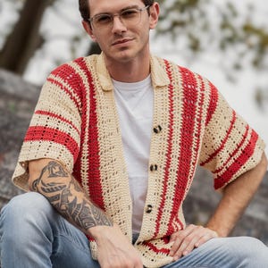 Può includere: Camicia all'uncinetto color crema con strisce verticali rosse. La camicia ha un colletto e tre bottoni scuri. La persona indossa una maglietta bianca e jeans blu. È un capo casual e fatto a mano, adatto alla stagione calda.