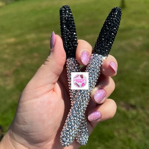 May include: Two rhinestone-covered makeup applicators. The handles are silver, and the tops are black. The applicators are held up in a peace sign gesture. The background is a green field.