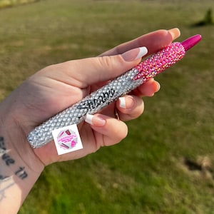 May include: A silver and pink bedazzled Sharpie pen. The pen is covered in rhinestones. The word "Shoobie" is written on the barrel. A small logo sticker is visible. The pen is held in a hand.