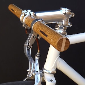 May include: A white bicycle with a wooden handlebar and silver brake lever. The handlebar is made of natural wood with a grain pattern. The brake lever is attached to the handlebar with a silver clamp.