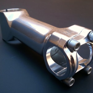 May include: Silver bicycle stem with a polished finish and multiple bolts.