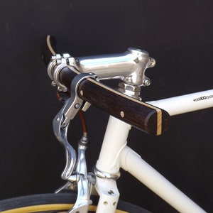 Può includere: Primo piano di un telaio di bicicletta bianco con un attacco manubrio e leve dei freni in argento lucido. Il manubrio è in legno scuro con accenti di legno chiaro. Lo pneumatico della bicicletta ha un fianco giallo.
