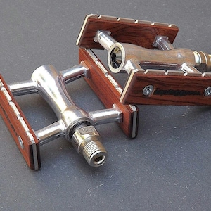 Puede incluir: Un par de pedales de bicicleta plateados con plataformas de madera. Las plataformas son de madera oscura con un patrón de veta y tienen un borde plateado.